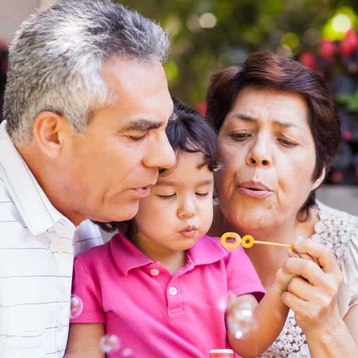 Older Hispanic couple sitting in a garden with a toddler blowing bubbles through a yellow bubble wand.