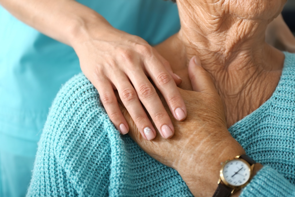 Older person's hand on shoulder with younger caregiver's hand on top in a caring manner.