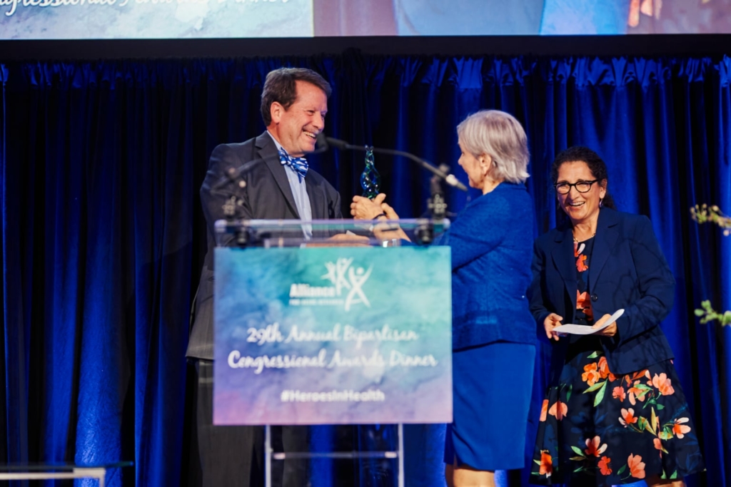 Dr. Robert Califf presents the Perennial Hero Award to Dr. Janet Woodcock on the stage.