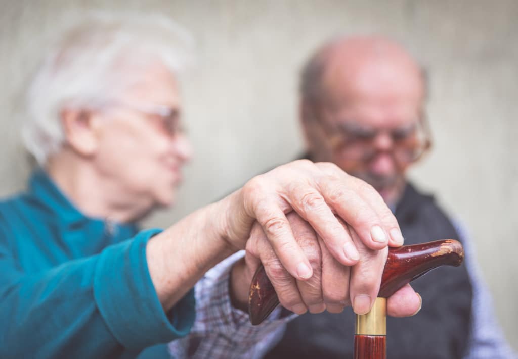 Two older adults with hands covering each other's hands on wooden cane.