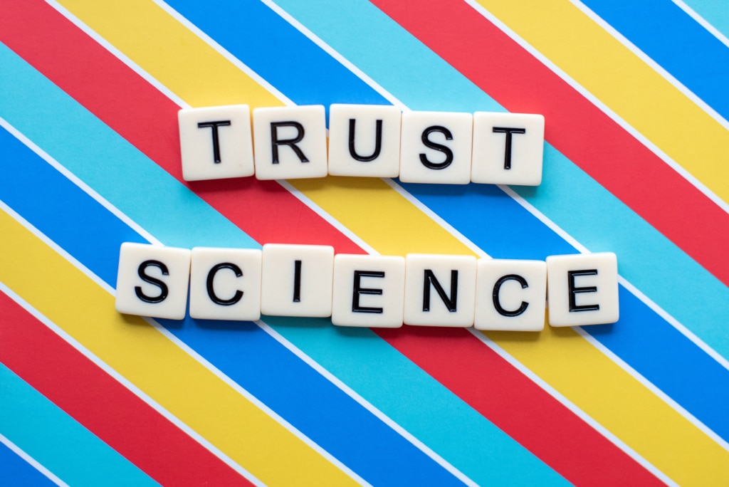 Black and white letter tiles spelling "Trust Science" in a striped colorful background.