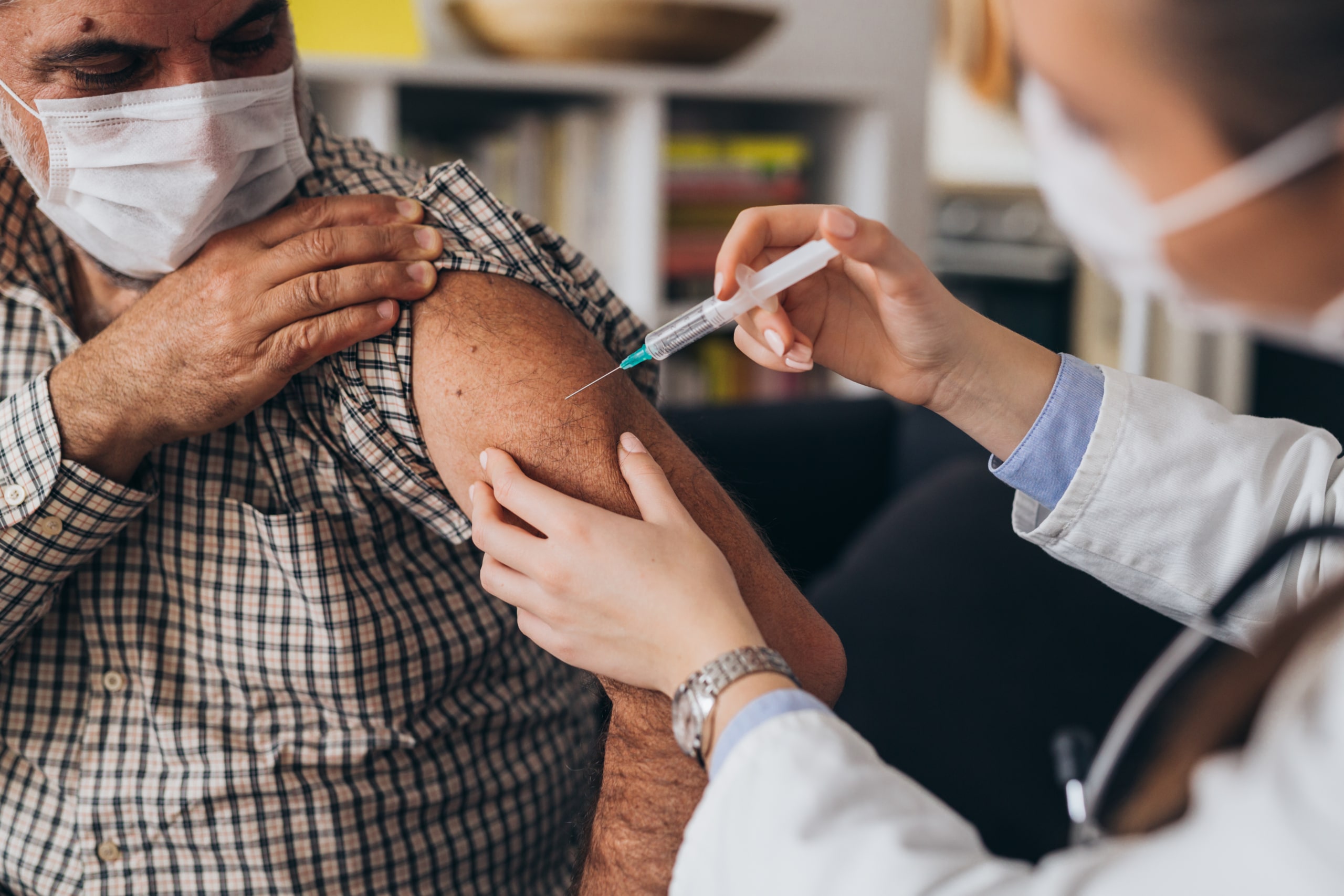 Masked older Hispanic male getting a vaccine administered by a physician in a white lab coat.
