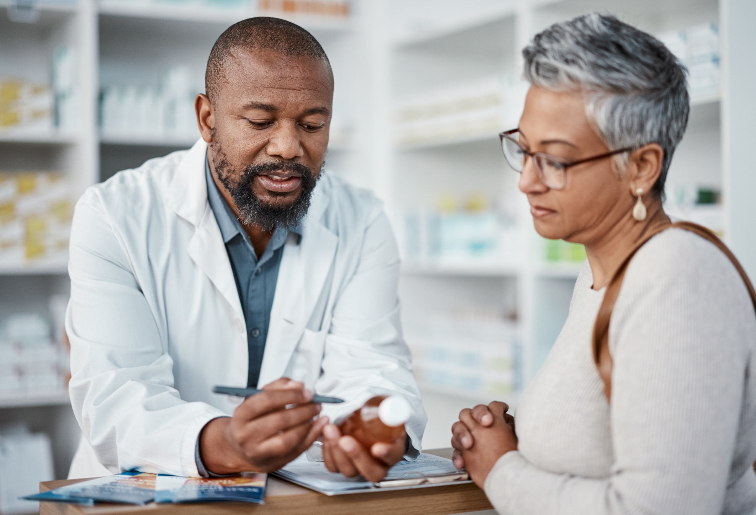 At the pharmacy counter, a black male pharmacist explains medication to a black senior woman.