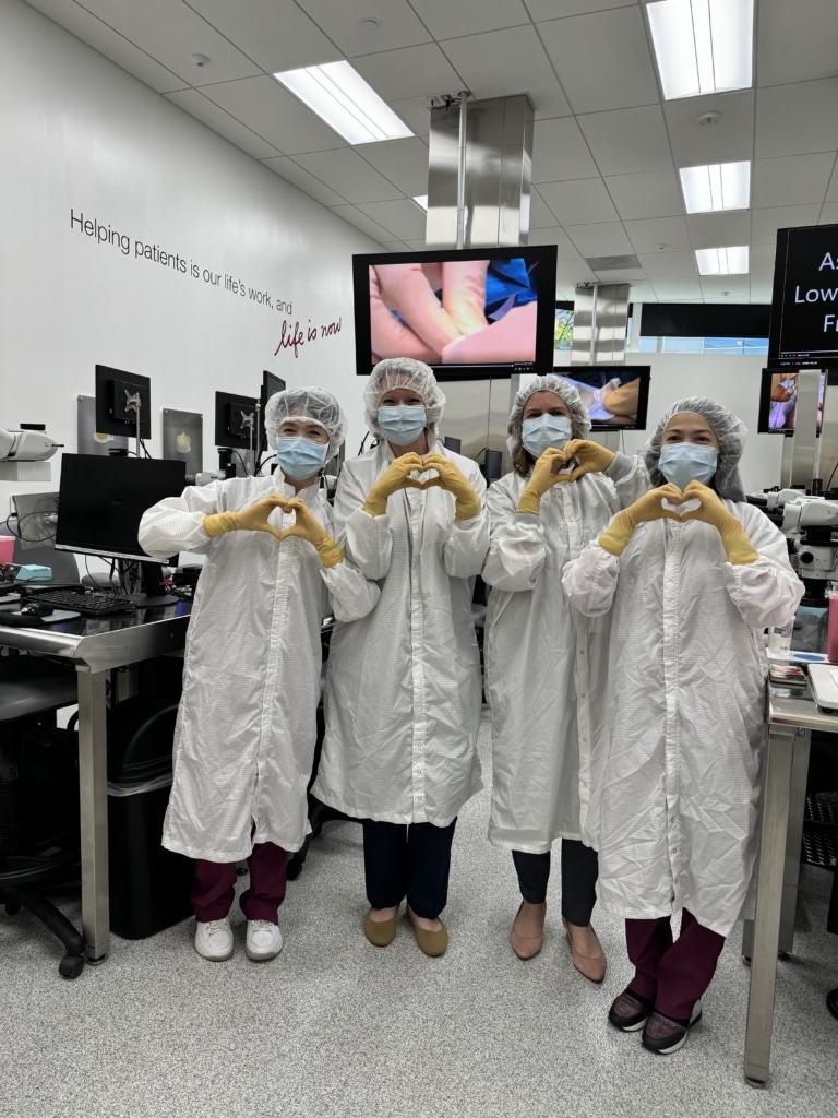 Wearing white lab coats and PPE and making heart signs with their hands, Alliance Vice President of Communications Katie Riley, and Alliance Senior Vice President of Health Education & Advocacy Lindsay Clarke (center) pose with expert heart valve sewers at Edwards Lifesciences.