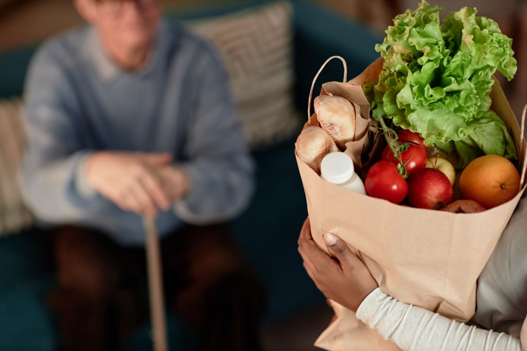 Person holding a brown grocery bag filled with nutritious food before an older adult with a cane.