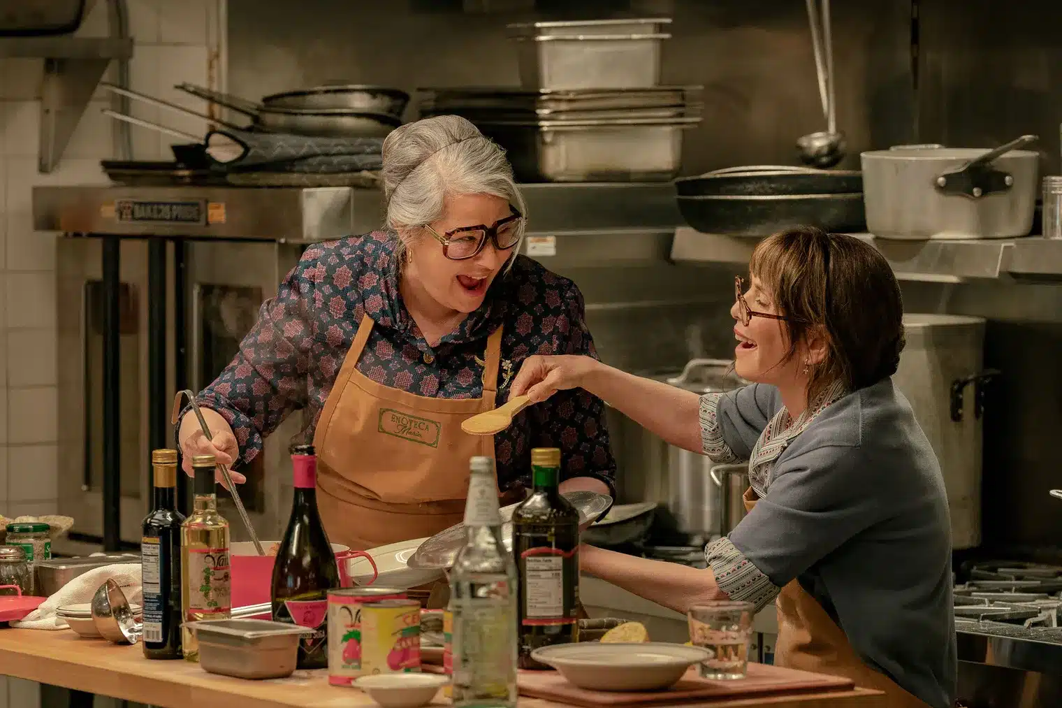 Two actresses in the Netflix movie "Nonnas" laughing over cooking in an industrial kitchen.