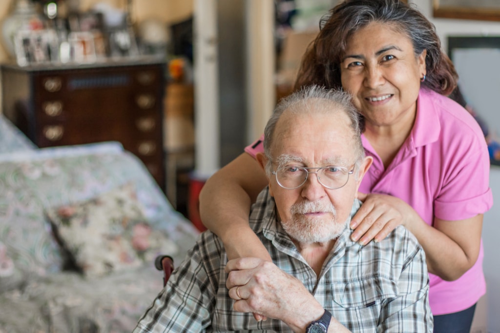 Older woman hugging a seated older man from behind while smiling in their home.
