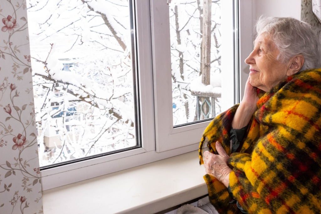 Older woman wrapped in a blanket looking out a window at the snow